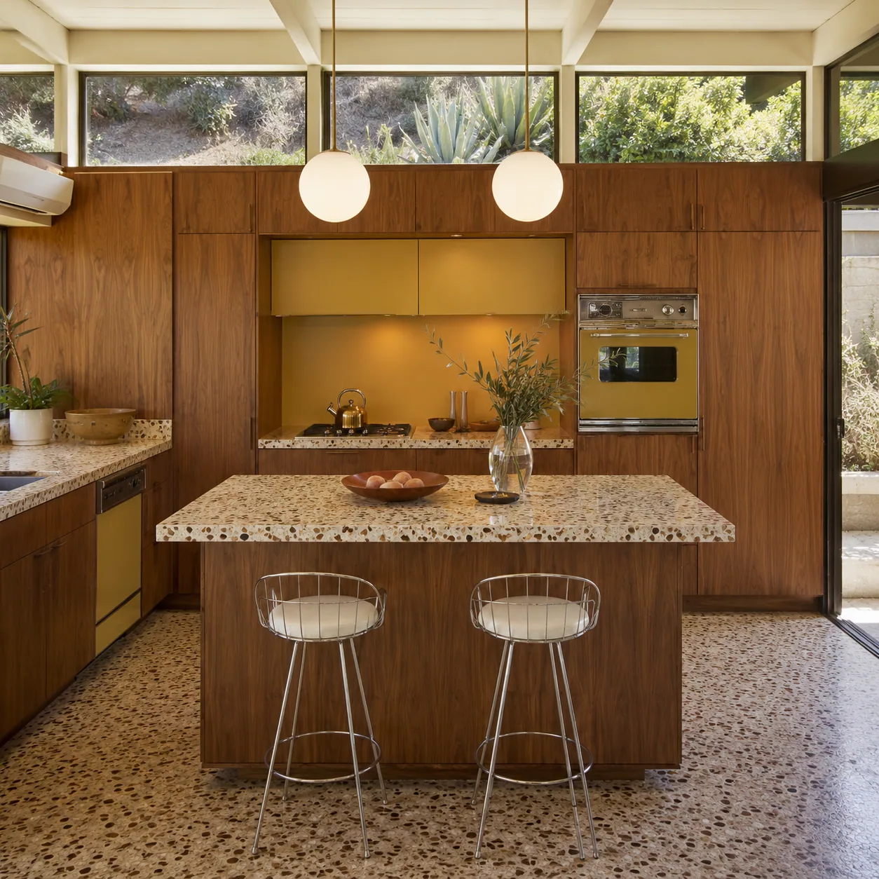 Square mid-century California kitchen with walnut cabinetry, terrazzo surfaces, a mustard niche, globe pendants and clerestory windows.