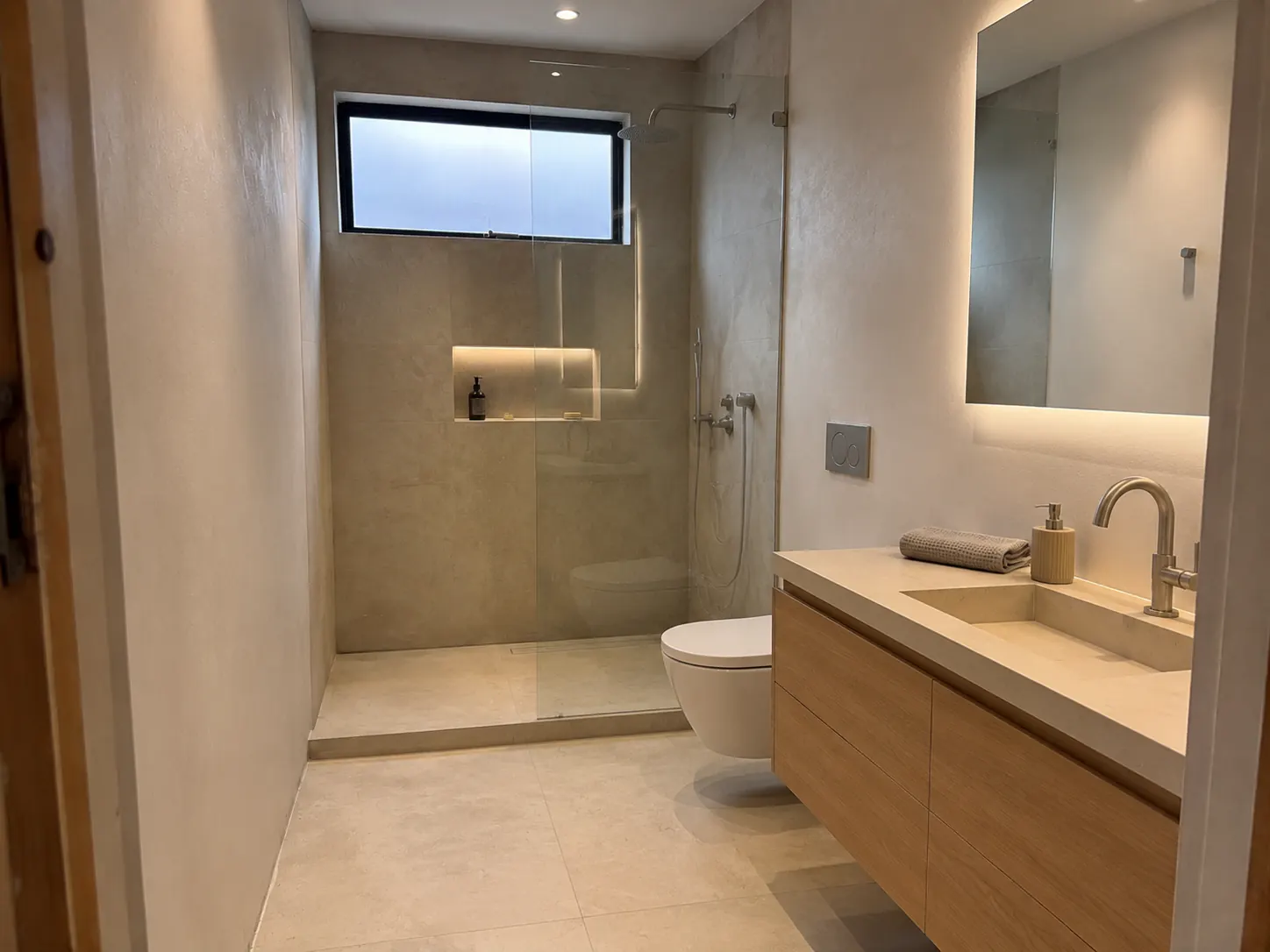 Restrained Modern Spa bathroom with warm limestone surfaces, floating oak vanity, frameless shower, and soft concealed lighting.