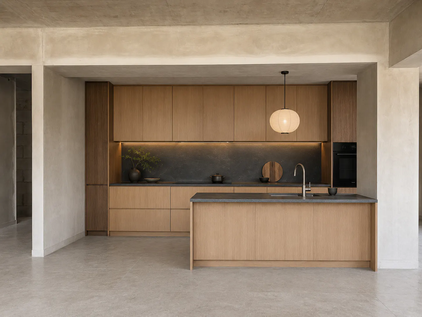 4:3 photograph of the same open-plan kitchen completed in restrained Japandi style with pale oak, charcoal stone, mineral plaster, and a compact peninsula